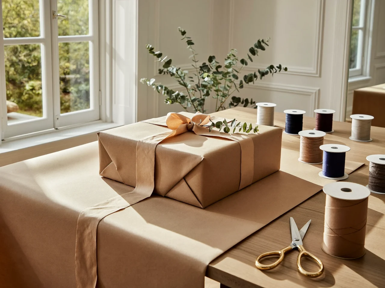 Heller Verpackungstisch am Fenster mit halb eingeschlagenem Geschenk, Kraftpapier, Bandspulen und Eukalyptuszweig